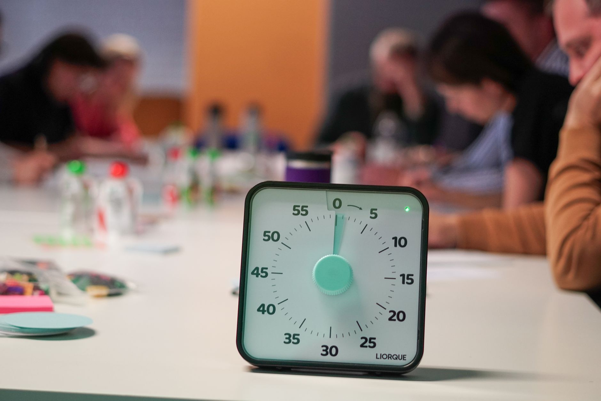 Small teams of adults at timed task stations in a bright hall with a facilitator holding a clipboard